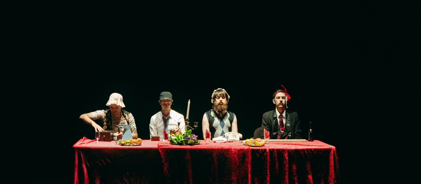 Das Ensemble STOPGAP sitzt auf der Bühne an einem roten Tisch