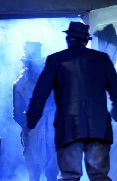 Das Bild zeigt den Rücken von einen Mann mit Hut, der zu einer Tür schaut, aus der eine Gestalt mit einem Hut im blauen Nebel hereintritt.