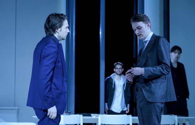 Alexandros Koutsoulis und Max Poerting in ihren Rollen stehen in Anzügen nebeneinander auf der Bühne. Im Hintergrund sind Jan Meeno Jürgens &amp; Janek Maudrich in ihren Rollen zu sehen.