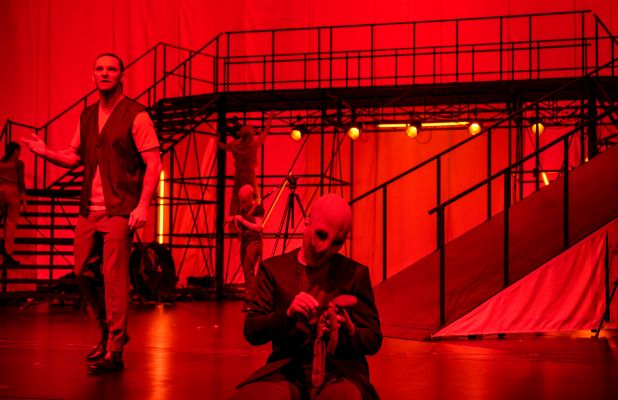 Der Erfinder und zwei Wesen mit unheimlichen Masken und Stoffpuppen in der Hand sind in rotes Licht getaucht.