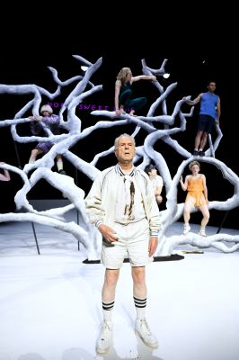 Hans Dieter Trayer in seiner Rolle steht ganz in weiß vorne auf der Bühne. Im Hintergrund ist das Ensemble sitzend auf dem weißen Baum zu sehen.