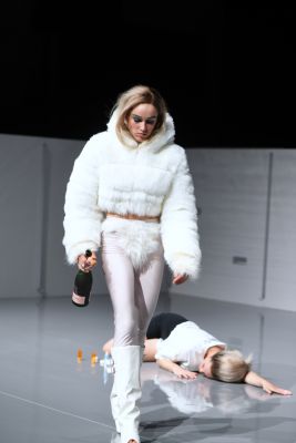 Die Rolle von Ruth Bohsung läuft über das Bühnenbild mit einer Flasche in der Hand und schaut auf den Boden. Die Rolle von Liv Stapelfeldt liegt im Hintergrund auf dem Boden.