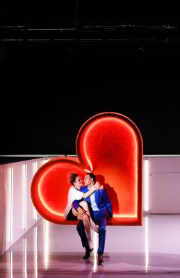 Alexandros Koutsoulis und Liv Stapelfeldt in ihren Rollen sitzen auf einer Bank in Herzform und schauen sich verliebt an.