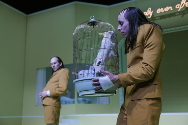 Lorenz Hochhuth in seiner Rolle steht hinten links und schaut nach vorne rechts zu der Rolle von Julian Gutmann. Dieser hält einen Käfig mit einem weißen Papagei darin.