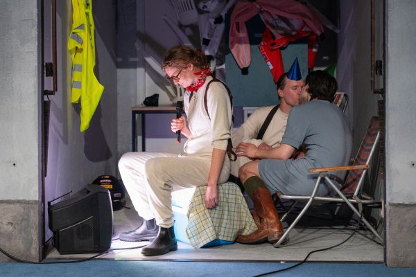 Die Rolle von Liv Stapelfeldt sitzt auf einer Kühlbox und schaut auf einen kleinen Fernseher. In ihrem Rücken sitzen die Rollen von Steffen Link und Jan Meeno Jürgens auf Gartenstühlen und küssen sich.