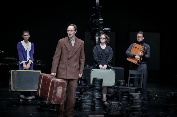 Die Rollen von Nina Noé Stehlin, Nils Thalmann, Maximiliane Haß und Jan Meeno Jürgens stehen nebeneinander. Sie sind umgeben von Stapeln an schwarzen Filmrollen, halten Koffer in ihren Händen und Armen und blicken nach vorne.
