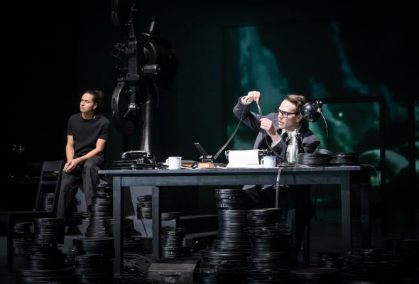 Die Rolle von Jawad Rajpoot sitzt links im Bild auf Stapeln von schwarzen Filmrollen. Rechts daneben sitzt die Rolle von Silas Breiding an einem Schreibtisch und inspiziert die Bilder einer Filmrolle genaustens. Die eingepackten Filmrollen stapeln sich auch um ihn herum.