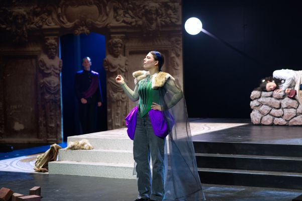 Steffen Link, Luise Deborah Daberkow (c) Gabriela Neeb Die Rolle von Steffen Link ist unscharf links im Hintergrund zu sehen und schaut nach vorne. Die Rolle von Luise Deborah Daberkow steht mittig auf der Bühne mit Hand in der Hüfte und gehobener Nasenspitze.