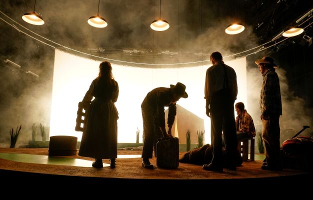 Es sind die Umrisse des Ensembles zu sehen, die mit dem Rücken zum Publikum stehen und in ein helles breitflächiges Licht im Hintergrund blicken.