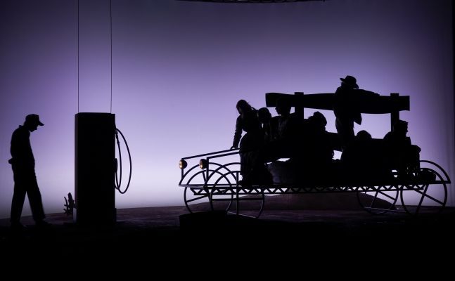 Es sind die Umrisse eines Wagons mit dem Ensemble darauf zu sehen. Der Hintergrund zeine eine lila Beleuchtung, die von unten nach oben dunkler wird.