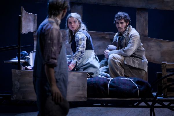 Die Rollen von Alexandros Koutsoulis steht unscharf im Vordergrund mit dem Rücken zum Publikum. Er blickt zu den Rollen von Anne Stein und Lukas Darnstädt, die in einem Wagon sitzend mitgenommen und erschöpft aussehen.