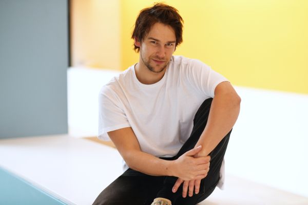 Portrait von Alexandros Koutsoulis sitzend vor einem gelben Hintergrund. Er blickt direkt in die Kamera.