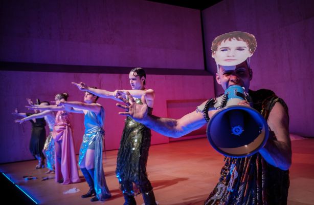 Das Ensemble tanzt auf der Bühne. Jonathan Müller in seiner Rolle steht rechts, trägt eine Caligula-Maske auf dem Kopf und schreit in ein Megafon.
