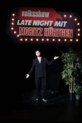 Moderator Moritz Hürtgen steht vor einem Schild mit der Aufschrift "volksshow: Late night mit Horitz Hürtgen"