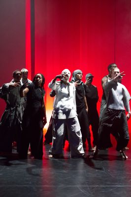 Eine Gruppe von Tänzer*innen steht in einer geballten Formation vor einem roten Vorhang.