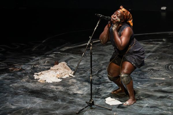 Ein Performerin reibt sich Lehm auf die Wangen und spricht in ein Mikrofon.