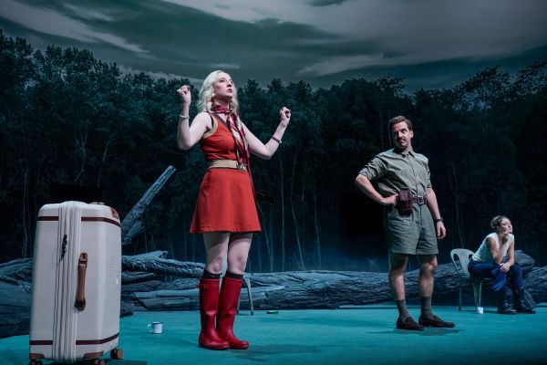 Carolin Hartmann steht entschlossen mit angewinkelt nach oben gehobenen Armen neben einem Koffer. Pascal Fligg steht rechts von ihr und schaut sie mit gehobener Augenbraue an. Lola Dockhorn sitzt neben ihm rauchend auf einem Stuhl und schaut gedankenverloren nach vorne.