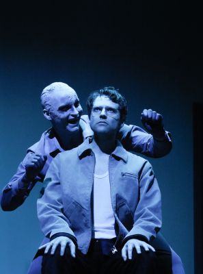 Anton Nürnberg als Krähe steht gestikulierend hinter Silas Breiding als Vater, der auf einem Stuhl sitzt.