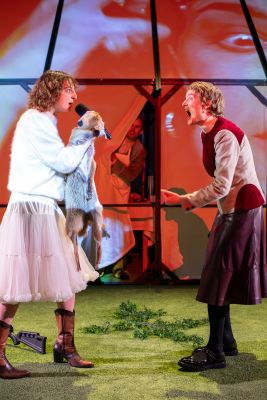 Zwei Schauspielerinnen streiten sich im Vordergrund, ein Spieler späht von hinten durch einen Vorhang.