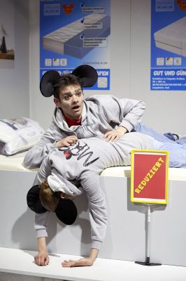 Zwei Personen liegen auf einer Matratze. Arme und Kopf von Luise Hart hängen über der Kante, Anton Andreew stützt sich auf ihrem Rücken auf. Beide tragen Mickey Mouse Ohren. Vor ihnen auf dem Boden steht ein "REDUZIERT" Schild.
