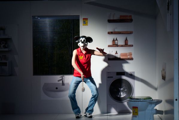 Luise Hart in Jeans und rotem Tanktop, bedruckt mit einem kleinen gelben Mickey Mouse Kopf, steht tänzerisch mit O-Beinen und hält eine Klobürste mittig vor sich. Auf dem Kopf trägt sie einen Mickey Mouse Kopf.