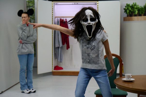 Vor einem geöffneten Kleiderschrank stehen zwei Darsteller:innen. Die Person vorne rechts im Fokus trägt ein graues Wonder Woman Shirt, sowie eine Ghostface Maske. Sie deutet nach links. Die Person hinten trägt Mickey Mouse Ohren, und kaut verängstigt auf den Nägeln.