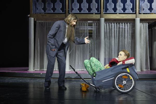 Katharina Brenner, gekleidet in übergroßem grau-karierten Anzug und mit angeklebtem Bart, redet gestikulierend auf Clara Kroneck ein, die mit einer grün gemusterten Hose und rot-weißer LEderjacke in einem Fahrrad-Anhänger sitzt