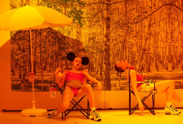 Luise Hart und Laura Fouquet liegen auf Campingstühlen. Luise trägt dabei Mickey Mouse Ohren und eine Bachtasche. An der Wand hinter ihnen klebt ein Bild eines Waldes. Links steht ein gelber Regenschirm. An den Möbeln hängen Preisschilder.