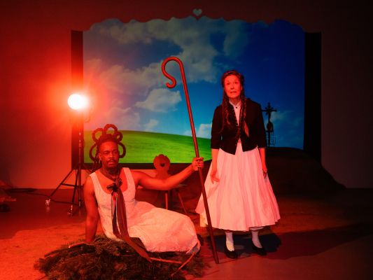 Challange Gumbodete sitzt in weißem Kleid auf Tannenzweigen. Seine Haare sind zu künstlerischen Zöpfen geflochten, die eine Blume um seinen Kopf formen. Um seinen Hals trägt er ein langes Band. In seiner linken Hand hält er einen Hirtenstock. Zu seiner rechten steht Anja Schweitzer in einem weißen Kleid in A-Linie, über dem sie eine schwarze Trachtenjacke trägt. Ihre Haare sind geflochten und hängen in Zöpfen über ihren Schultern herab. Hinter ihnen ist blauer Himmel, Gras, sowie ein Gipfelkreuz zu sehen.