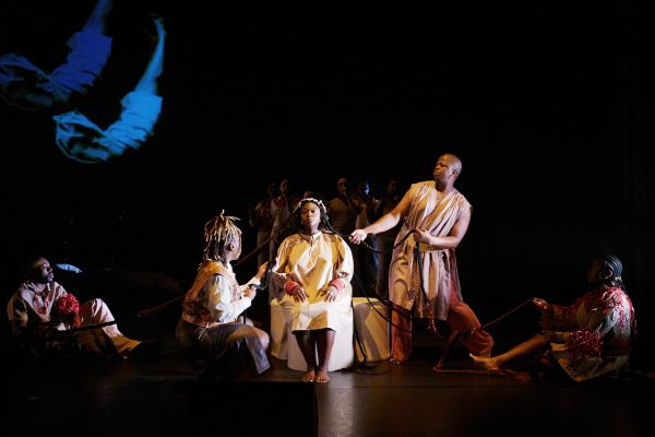 Eine Darstellerin sitzt mittig mit dem Blick nach vorne. Die anderen Ensemblemitglieder halten ihre langen Zöpfe.