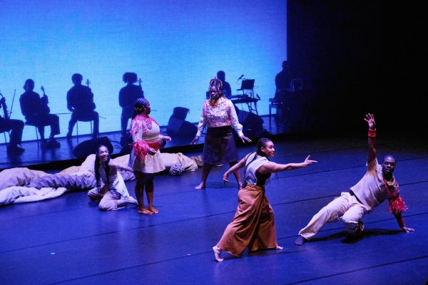 Ensemblemitglieder in tänzerischen Bewegungen. Hinter ihnen sind Schatten von Musiker:innen hinter einem transparenten Vorhang sichtbar.