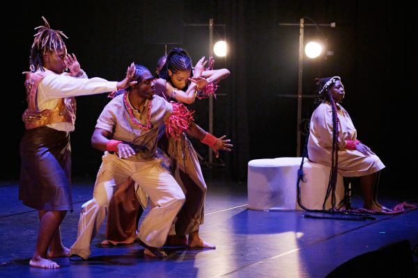 Auf der linken Bildhälfte sind mehrere Darsteller:innen in tanzenden Posen zu sehen. Rechts sitzt eine Frau mit Blick zum Publikum.