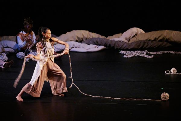 Ein Ensemblemitglied im Hintergrund flicht eine Schnur, während ein weiteres Ensemblemitglied eine bereits geflochtene Schnur schwingt.