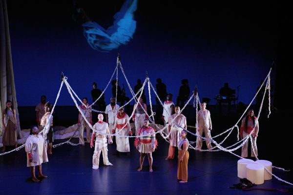 Das Ensemble steht verteilt auf der Bühne. Ihre Blick sind nach vorne gerichtet. Von oben herab hängen geflochtene Seile.