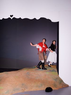 Anja Schweitzer in weißem Kleid und Trachtenjacke zieht Victor Calero an der Hand. Er blickt zurück und streckt seine andere Hand winkend aus. Über seinen Schultern hat er ein rotes Tuch geknotet.