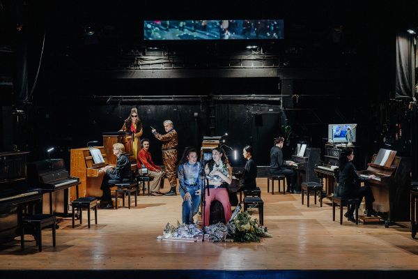 Acht Klaviere verteilt auf der Bühne. Vier Pianist:innen spielen. Zudem sind weitere Darsteller:innen auf der Bühne verteilt.