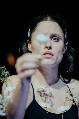 Eine Schauspielerin hält eine Blume in der Hand, sodass ihr linkes Auge verdeckt ist. Unter ihren Schlüsselbeinen, sowie an ihren Armen sind mit Pflastern weitere Blumen befestigt. Ihr Blick ist sehnsuchtsvoll.