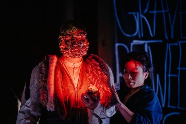 Ein Darsteller in rotem Licht. Sein Gesicht rot vernarbt und verdreifacht. Neben ihm steht eine Frau die seinen Pelz anfasst.