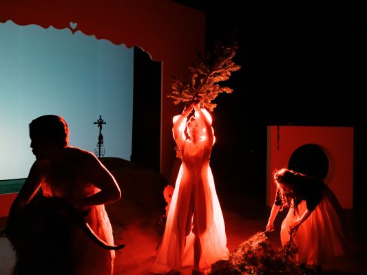 Drei Darsteller:innen sind in einer rot beleuchteten Szene abgebildet. Elisa Lynn Dillier in der Mitte hält einen großen Tannenzweig in die Höhe. Das Licht lässt ihr weißes Kleid hell erstrahlen. Rechts neben ihr sammelt Anja Schweitzer weitere Zweige auf.