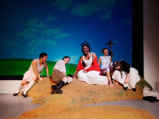 Fünf Ensemlemitglieder seitzen lachend auf einem Hügel. Alle tragen weiße Kleider, Röcke oder Hemden. Hinter ihnen tut sich eine Landschaft mit grünen Wiesen und blau bewölktem Himmel, sowie einem Gipfelkreuz auf.