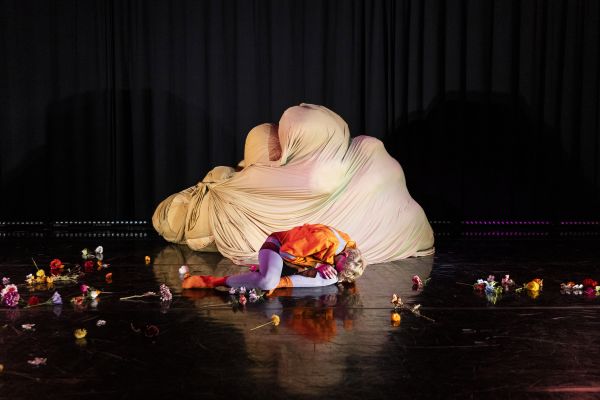 Johannes Hegemann liegt zusammengekauert vor einem weißen Stoffhaufen. Er trägt knallige Klamotten in lila und orange. Um ihn herum liegen auf der Bühne verteilt eine Vielzahl an bunten Blumen.