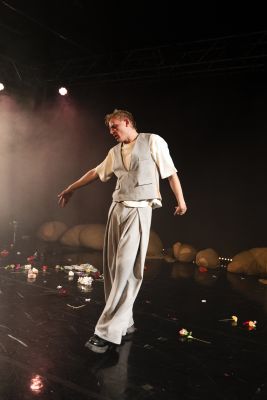 Ein Foto von Johannes Hegemann in einer gehend-tänzerischen Bewegung. er trägt eine hellgraue weitgeschnittene Anzughose, eine Weste und darunter ein weißes T-Shirt. Auf dem spiegelnden Boden sind einige Blumen verteilt. Im Hintergrund sind Stoffhaufen zu sehen.
