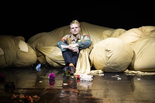 Johannes Hegemann sitzt vor einem großen Stoffballen auf schwarzem, spiegelnden Boden, umringt von vereinzelten bunten Blumen