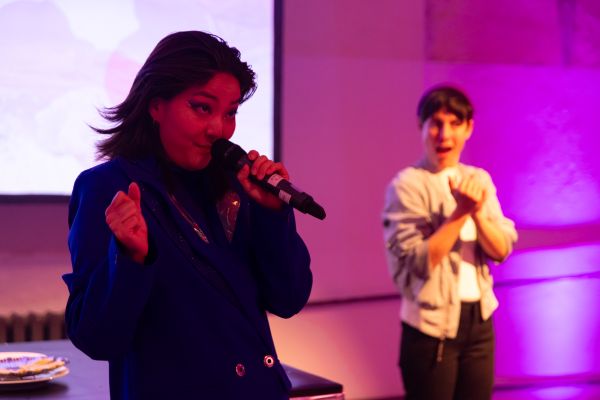 Eine Frau mit schwarzen Haaren spricht in ein Mikrofon. Rechts ist im Hintergrund eine weitere Person mit erstauntem Blick zu sehen.