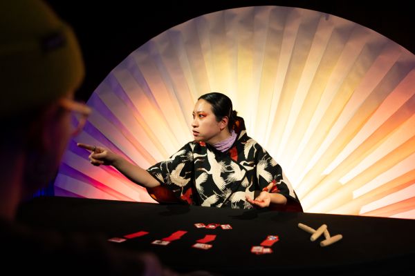 Olivia Hyunsin Kim sitzt in einem mit Pelikanen bedruckten Kimono und auffälligem orangen Augenmakeup vor einem großen weißen Fächer an einem Tisch und deutet auf eine Person außerhalb des Bildes. Vor ihr liegen etliche rote Karten mit Motiven.