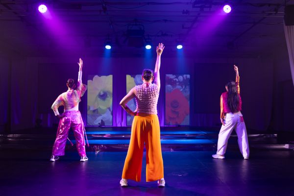 Drei Darstellerinnen mit breit geschnittenen farbigen Hosen stehen breitbeinig mit dem Rücken zum Publikum. Dabei haben sie jeweils den rechten Arm gerade nach oben gestreckt und den linken Arm in die Hüfte gestützt. Im Hintergrund sind drei hochkant ausgerichtete Bildschirme mit Blumen sichtbar.