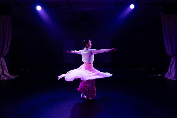 Eine Darstellerin dreht sich mit ausgestreckten Armen. Dabei fliegt ihr luftiger Rock tellerartig hoch. Unter ihrem Rock trägt sie eine breite rosa Hose. Sie wird von zwei Scheinwerfern blau beleuchtet.