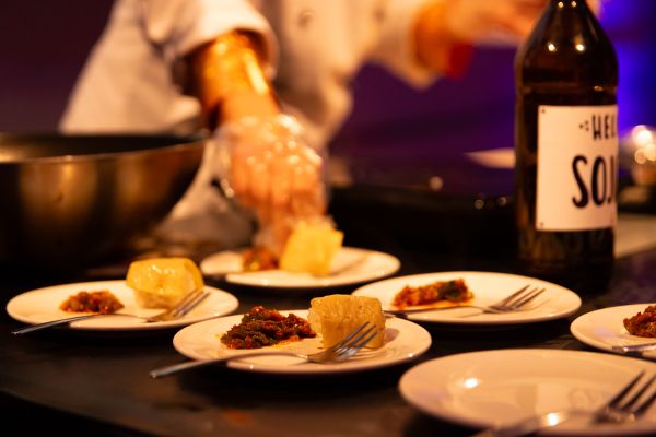 Eine Person in Kochjacke richtet mit Plastikhandschuhen Mahlzeiten auf Tellern an. Dabei sind die Teller im Fokus.