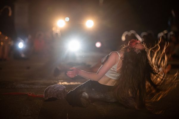 Eine am Boden kniende Frau, deren Haare in Bewegung schwingen. Mehrere Lichtquellen strahlen wie Taschenlampen in Richtung Kamera.