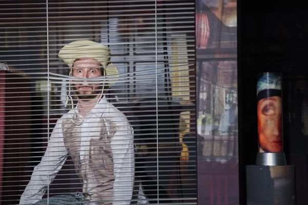 Ein Schauspieler mit Strohhut blickt nach vorne durch Jalousien hindurch.